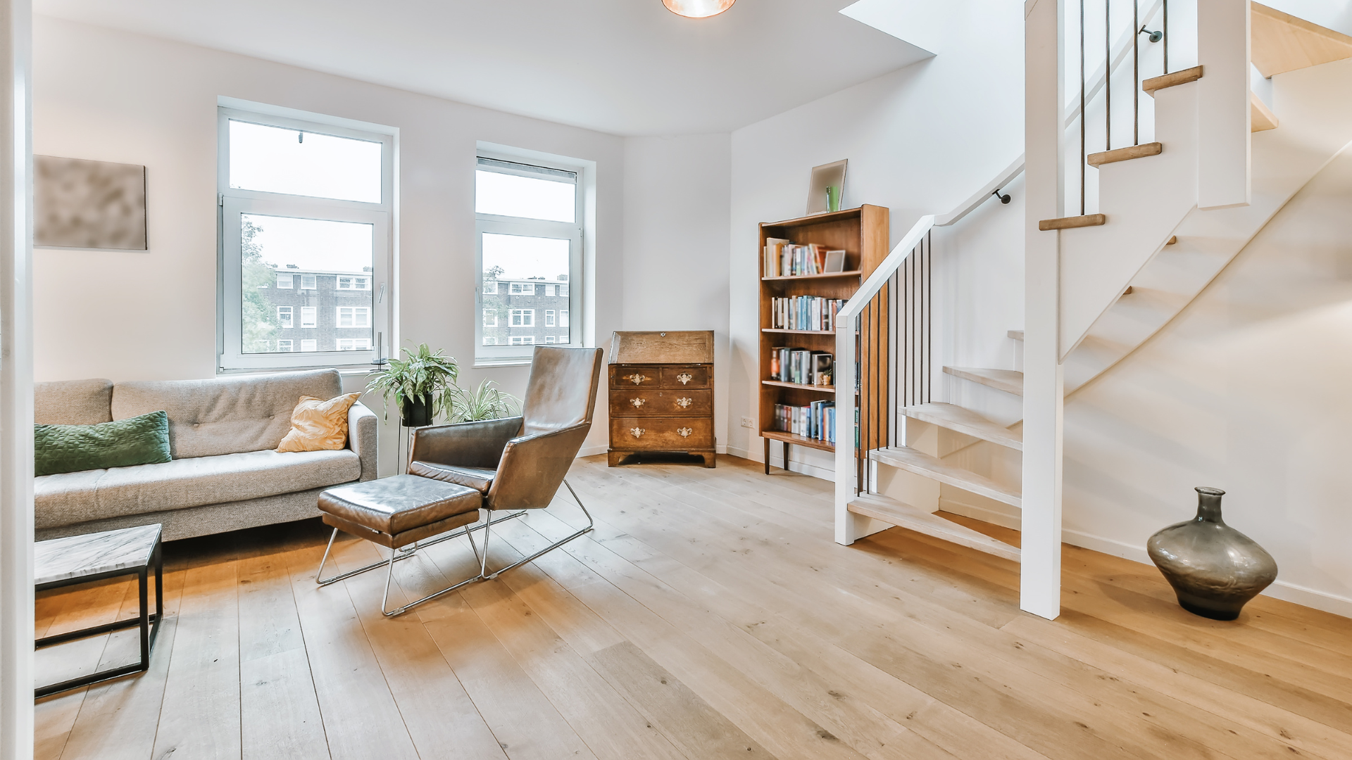 Bright living room with large windows, gray sofa, woven chairs, wooden floor, and staircase