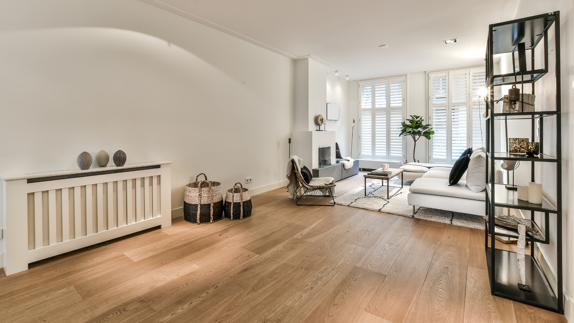 Bright minimalist living room with wood floors, white sofa, shelving, and a large window.