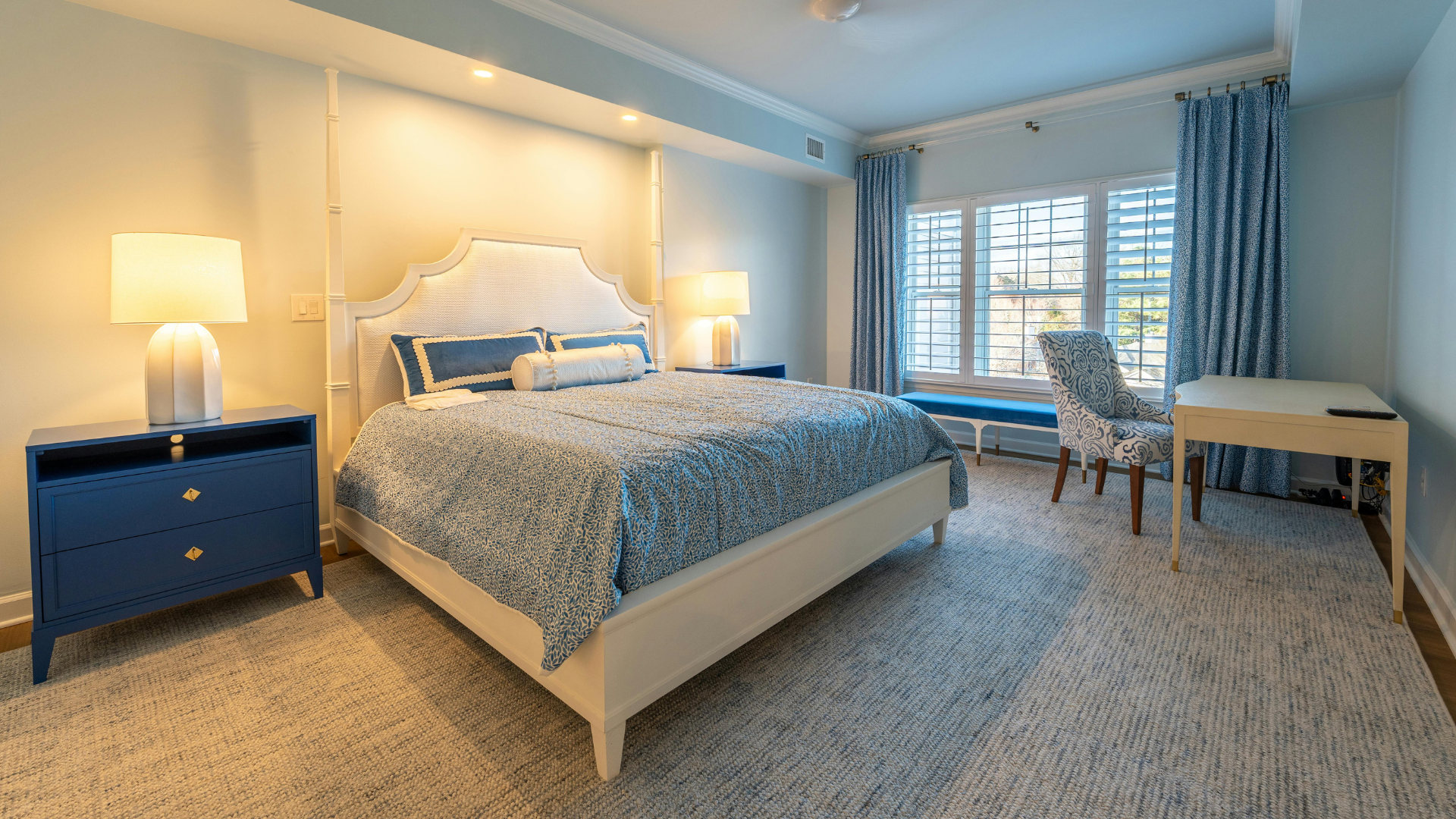 Bright bedroom with a blue bed, nightstands, desk, and large window with shutters