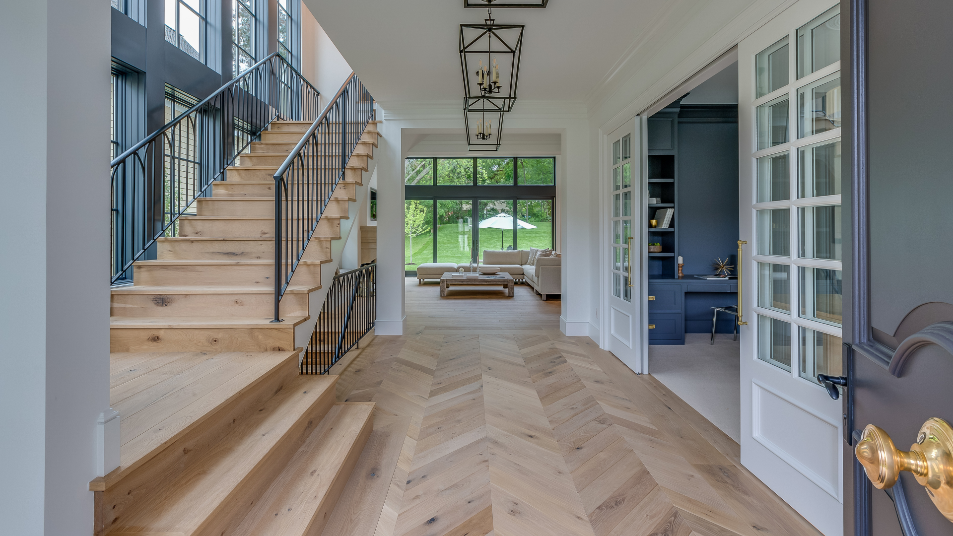 Bright foyer with herringbone wood floor, staircase, glass doors, and a view to a sunlit living area