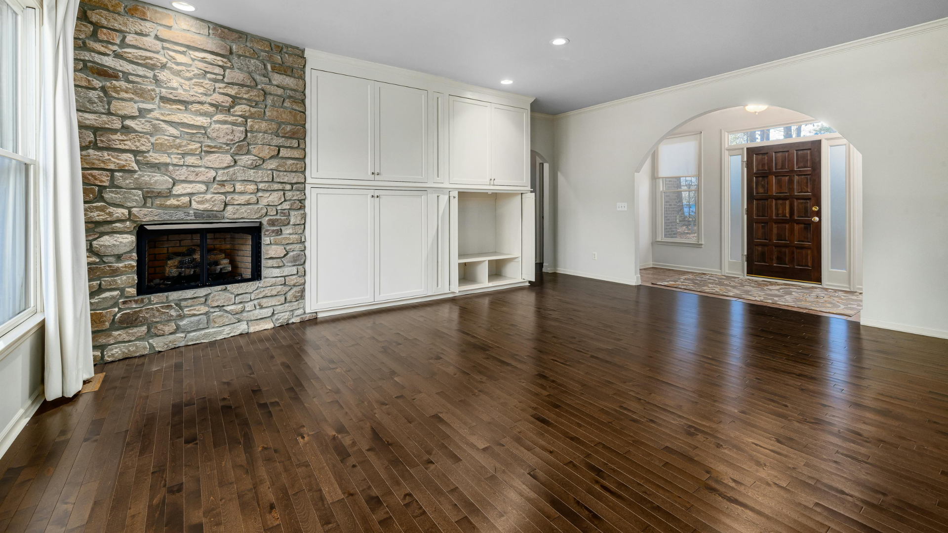 Spacious living room with hardwood floors, stone fireplace, white built-ins, and arched doorway to entry hall