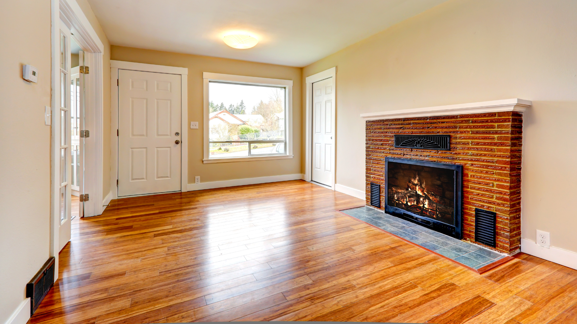 Empty living room with hardwood floors, beige walls, a window, and a brick fireplace.