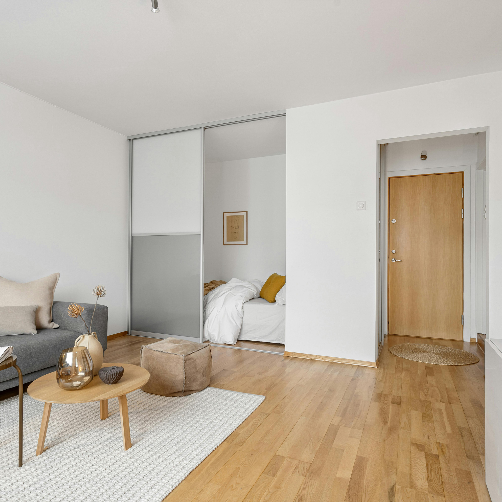 Minimalist living room with gray sofa, round wooden table, and a small bedroom nook behind sliding doors.