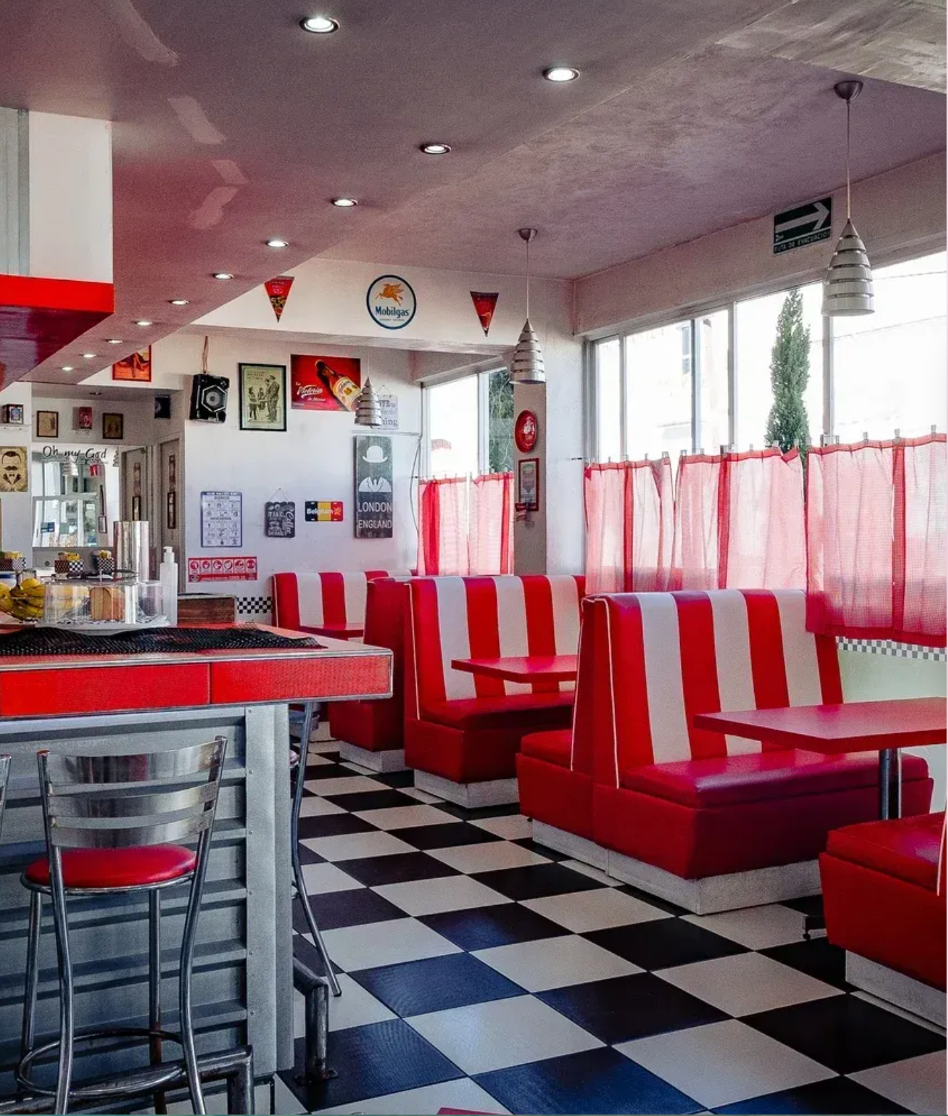 Interior de restaurante en rojo y blanco con asientos tipo cabina, suelo a cuadros y grandes ventanales.