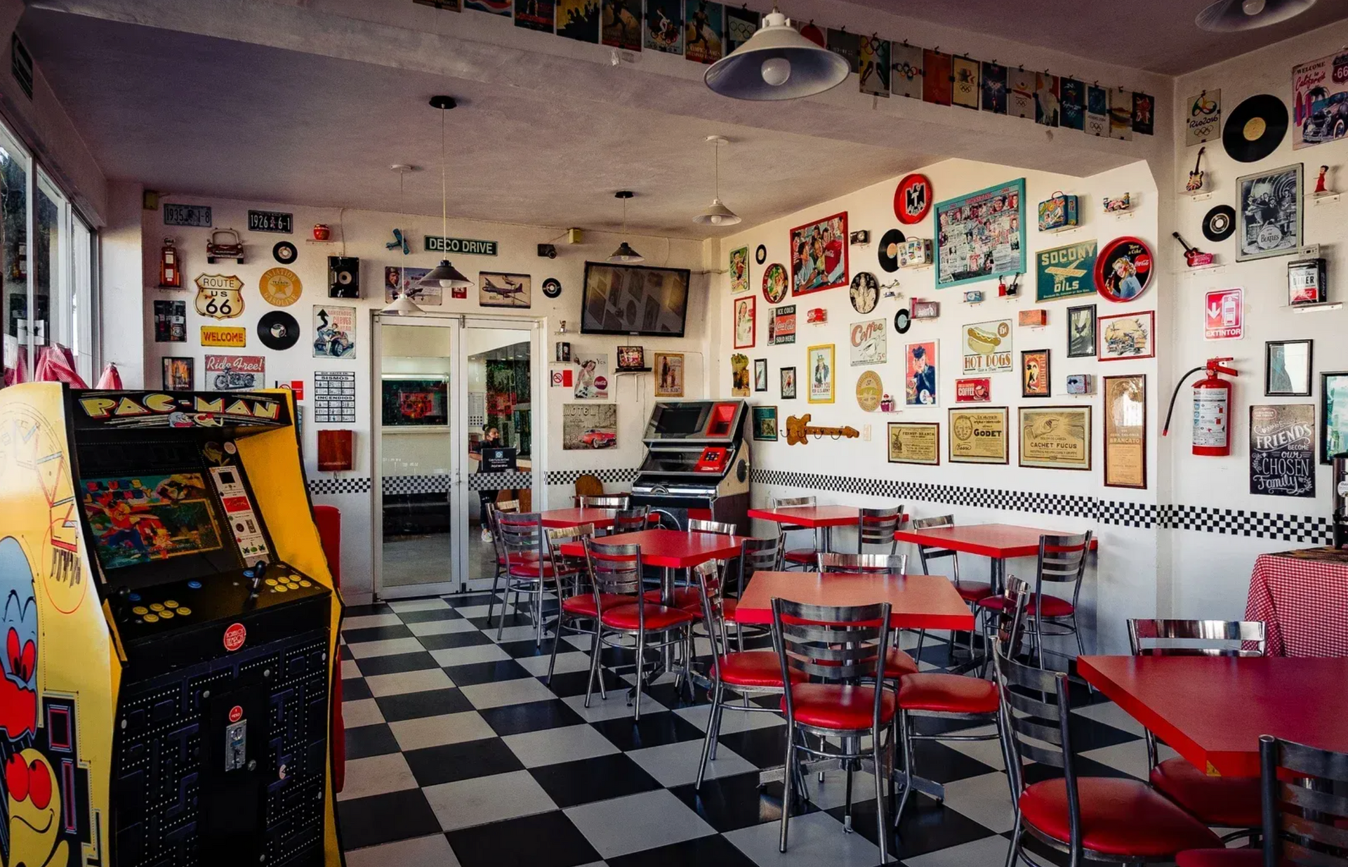 Restaurante retro con taburetes rojos, suelo a cuadros y paredes cubiertas de letreros y objetos de colección antiguos.