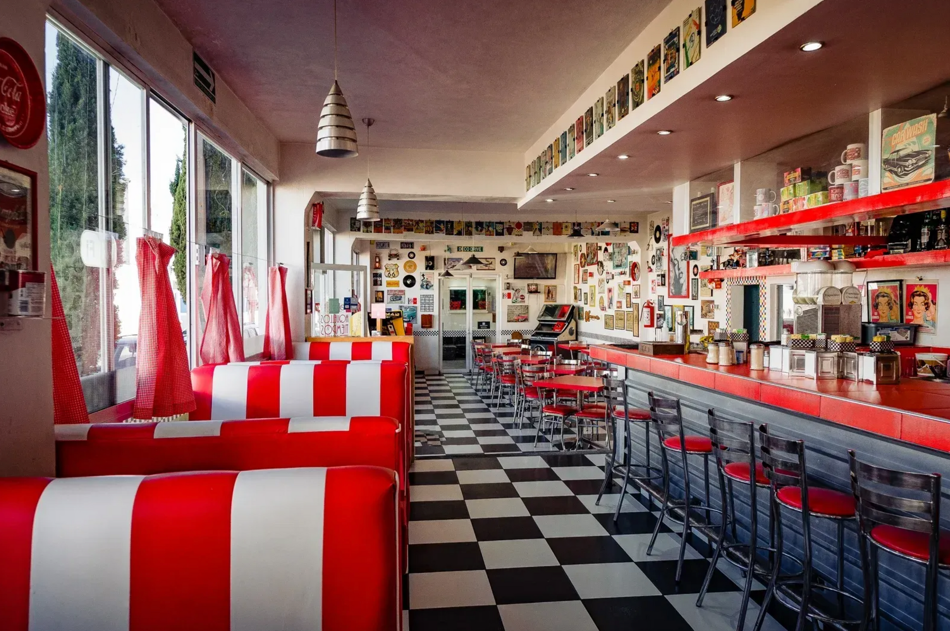 Interior de cafetería en rojo y blanco con suelo a cuadros, taburetes rojos y cabinas a lo largo de una larga barra.