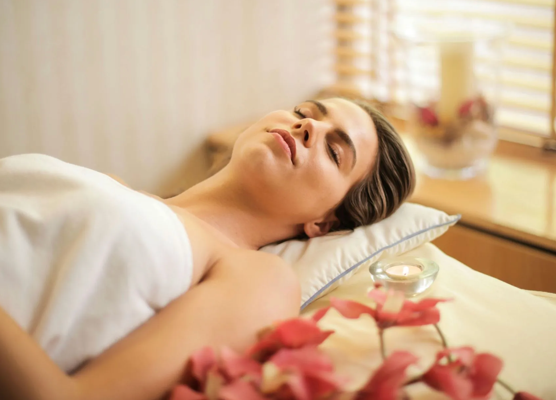 Mujer tumbada en una camilla de masaje de spa con los ojos cerrados, rodeada de flores y velas.