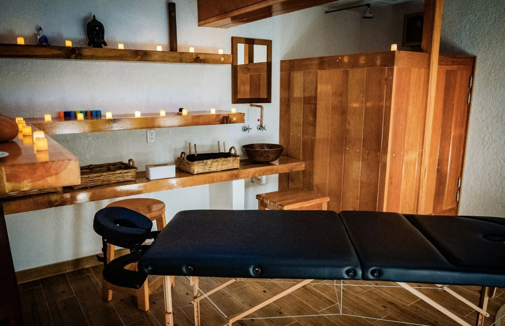 Sala de masajes con camilla de tratamiento, estantes de madera, velas y luz ambiental cálida.