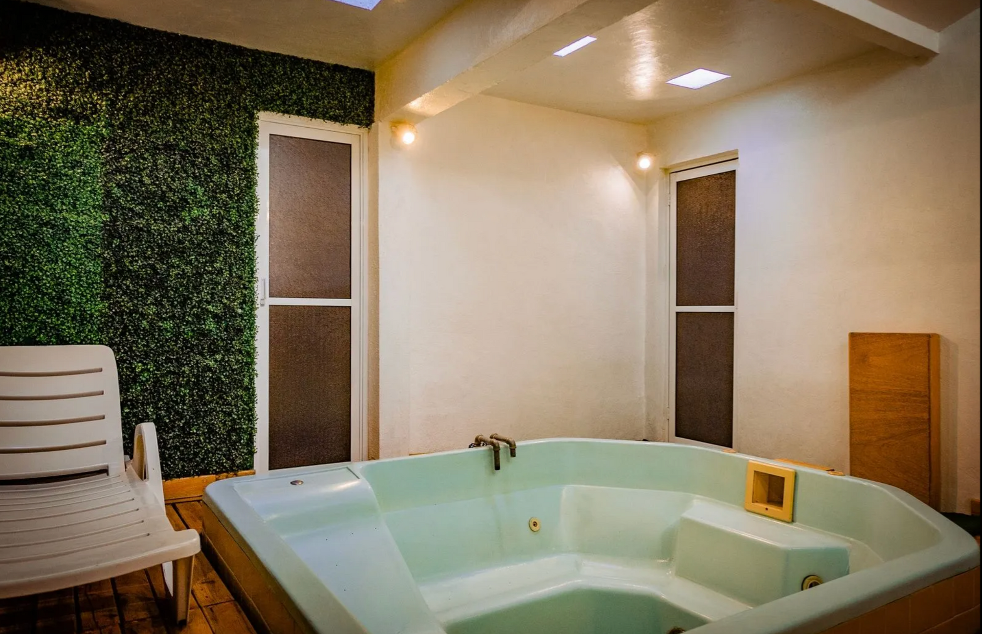 Sala de spa moderna con bañera de hidromasaje blanca, banco de madera, luces empotradas y pared vegetal.