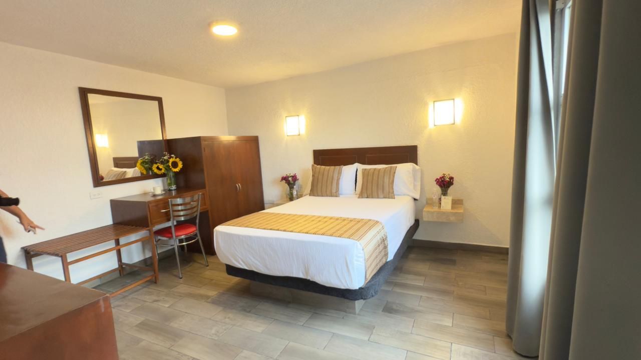 Habitación luminosa con cama, escritorio, espejo y cortinas que van del suelo al techo a la derecha.