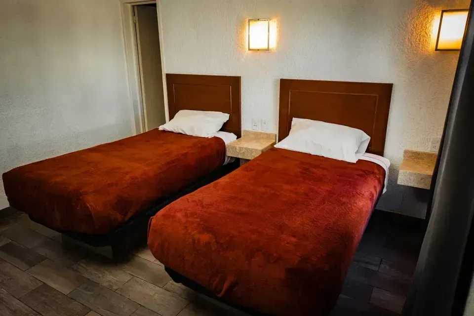 Dos camas individuales con mantas marrones en una sencilla habitación de hotel, iluminada por lámparas de pared.