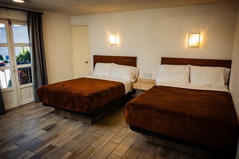 Habitación de hotel con dos camas, mantas marrones, apliques de pared y una ventana a la izquierda.