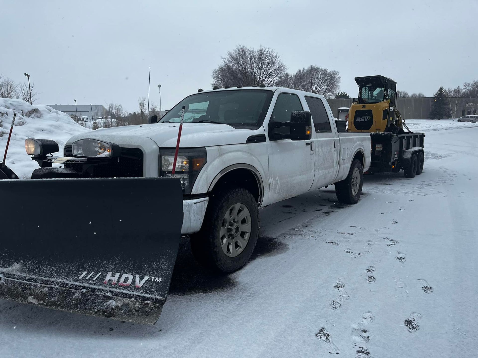 White pickup truck with snowplow attached pulling a trailer with a skid steer on a snowy surface.