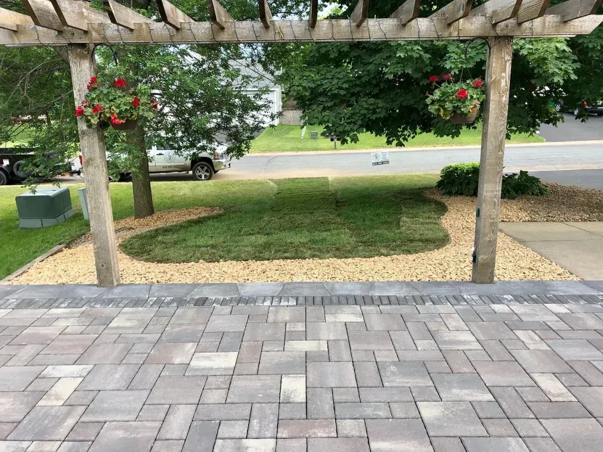 A patio leads to a small lawn bordered by tan gravel under a wooden pergola with hanging red flowers.