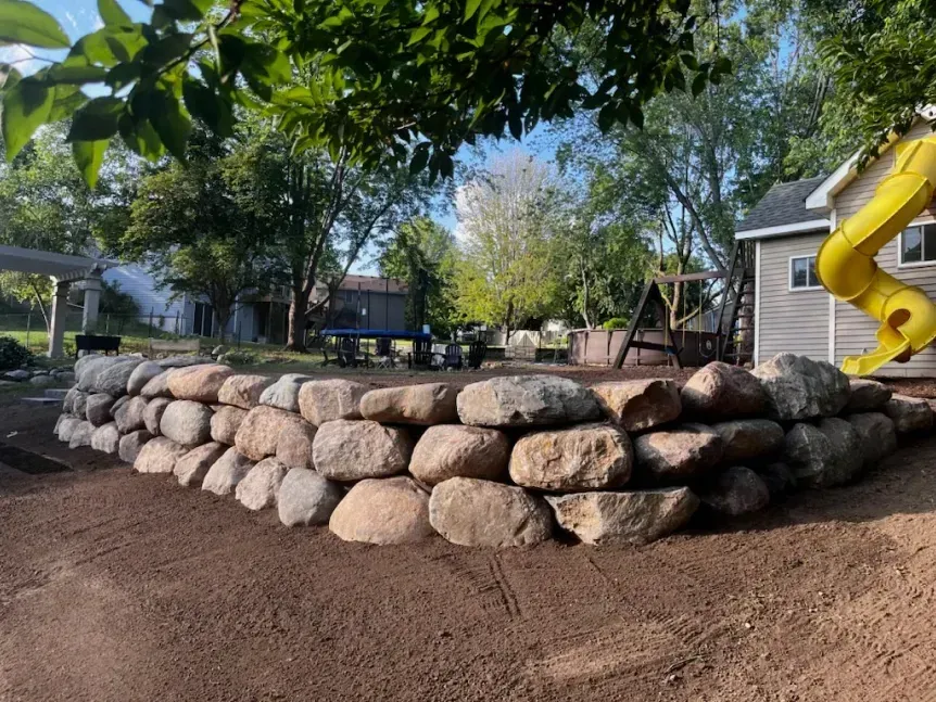 A retaining wall made of large, rounded stones surrounds a playground area with a yellow slide and swing set.
