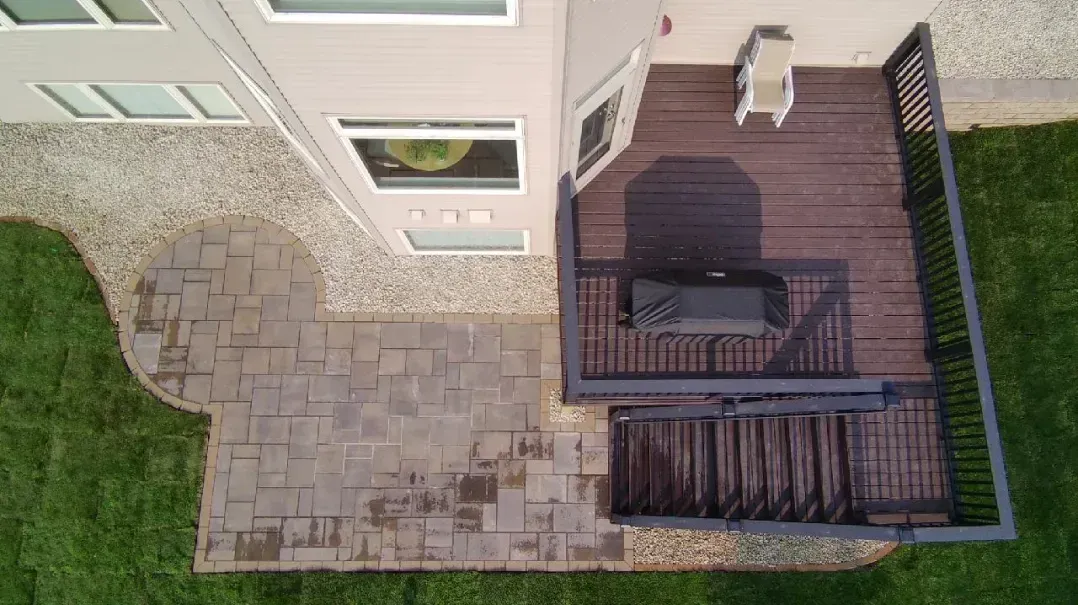 Overhead view of a house with a wooden deck and a stone patio. The deck has a grill, and the patio meets a green lawn.