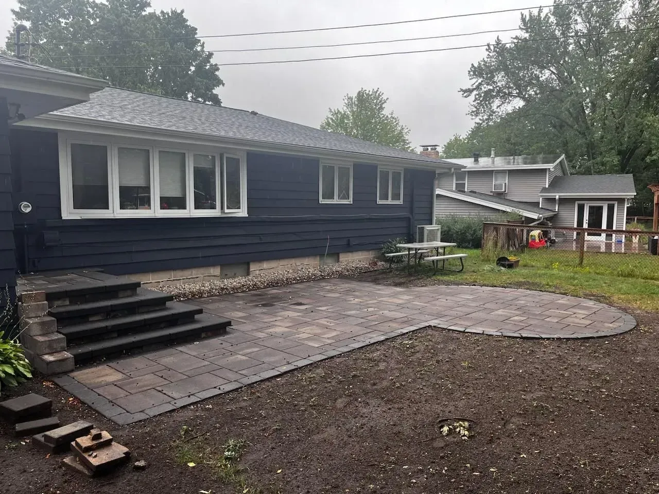 A newly constructed paver patio outside a dark blue house with steps leading to it; a picnic table is visible on the grass.