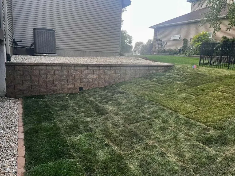 A backyard with a stone retaining wall creating a patio area next to a house. New sod laid on the lawn.