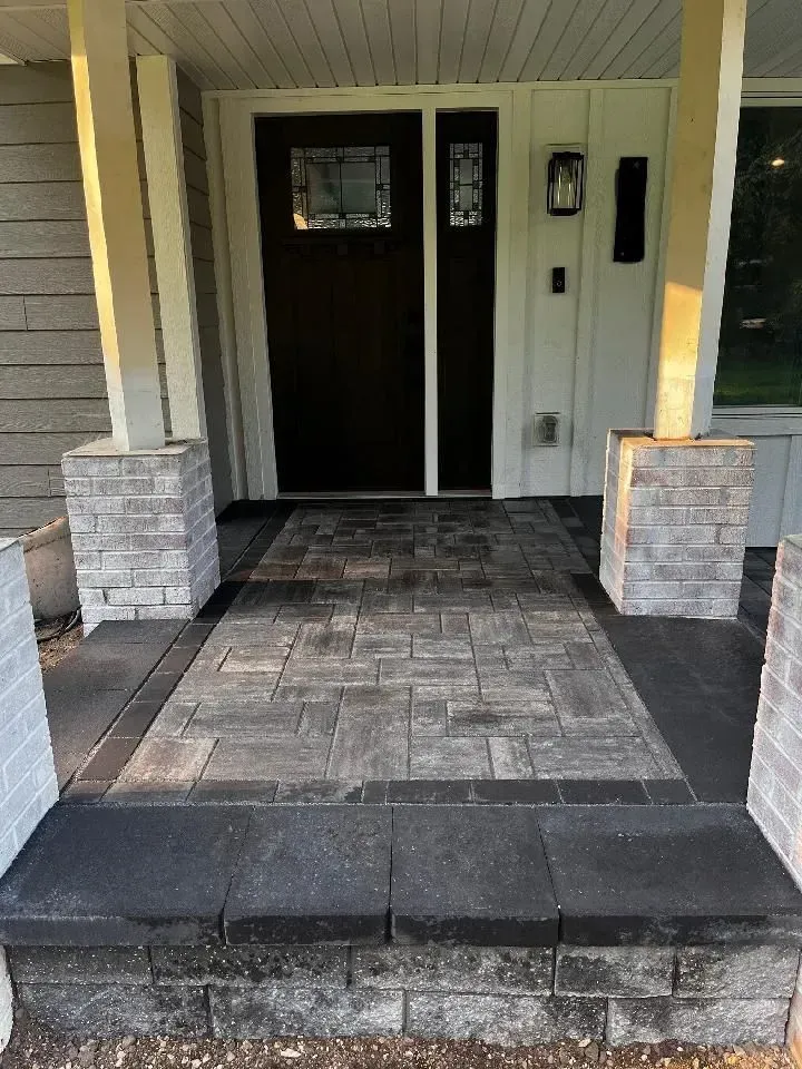 A front porch with brick pavers and pillars. The door is dark, flanked by white trim and gray brick.