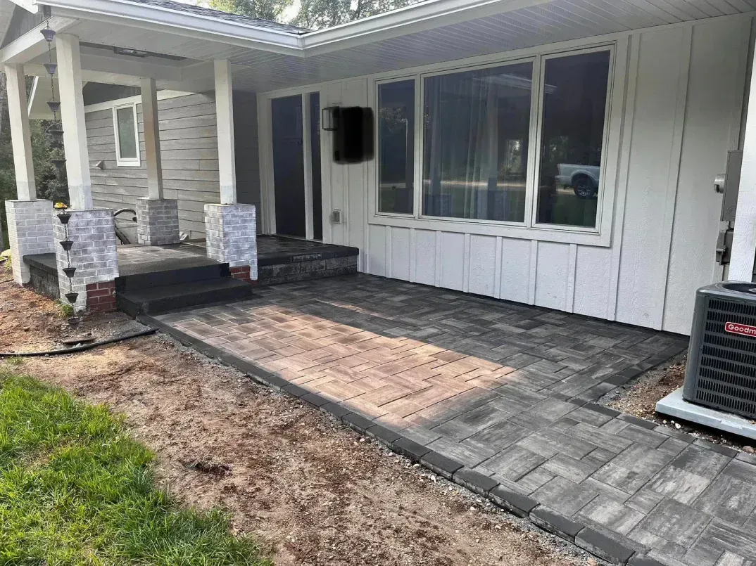 A house exterior with a paved walkway and patio. The house is white with large windows, and the walkway is made of 