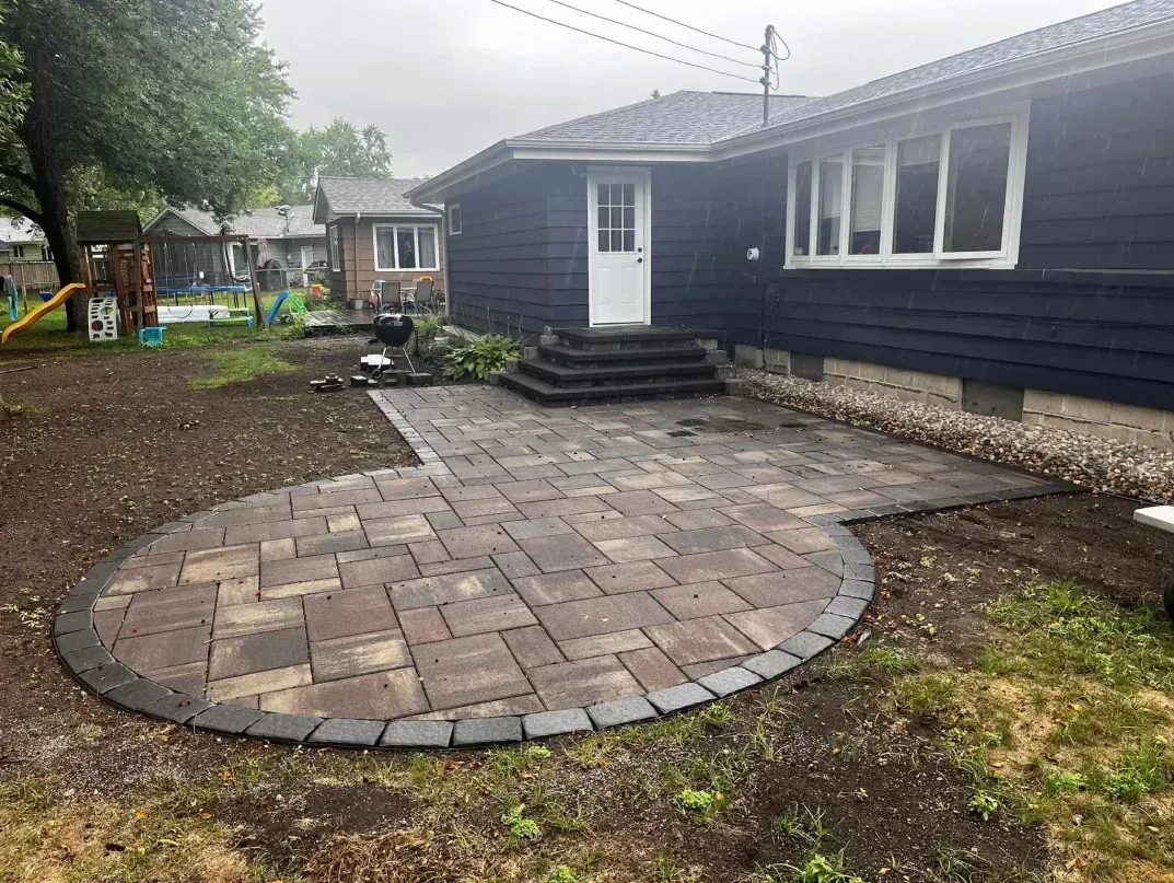 Brick patio in backyard, next to dark blue house with white door and steps. Overcast day.