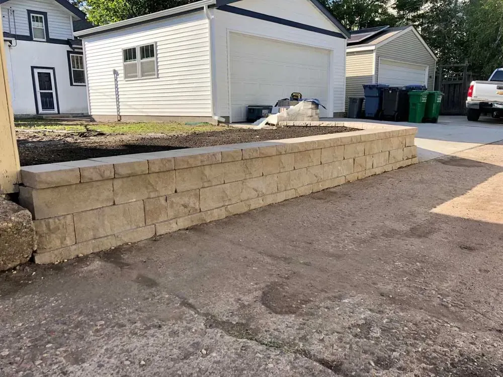 A tan retaining wall in front of a garage and driveway, with a small patch of dirt behind it.