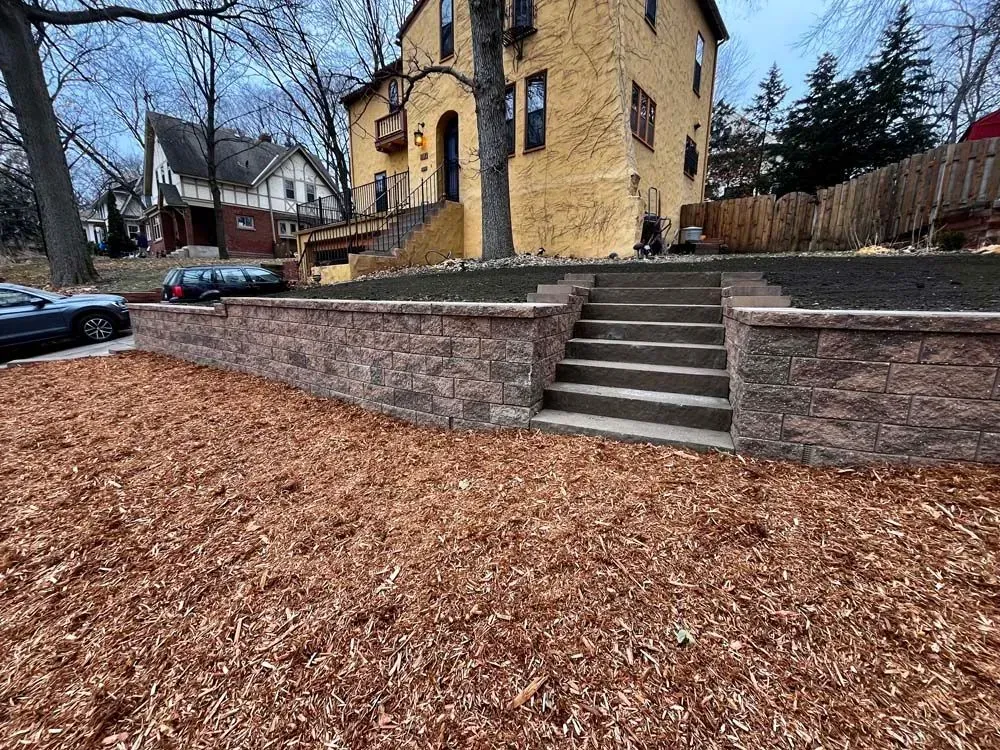 A two-tiered brick retaining wall with steps leading up to a yellow stucco house; a car parked on the street.