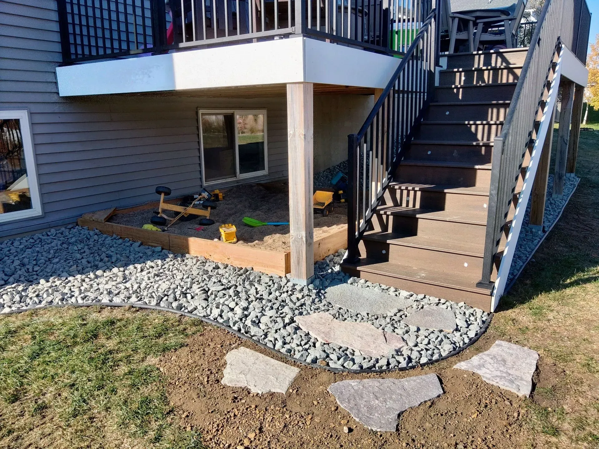 A backyard with a deck and stairs. Gravel borders a path of stone pavers leading to the stairs. Below the deck is a sandbox.