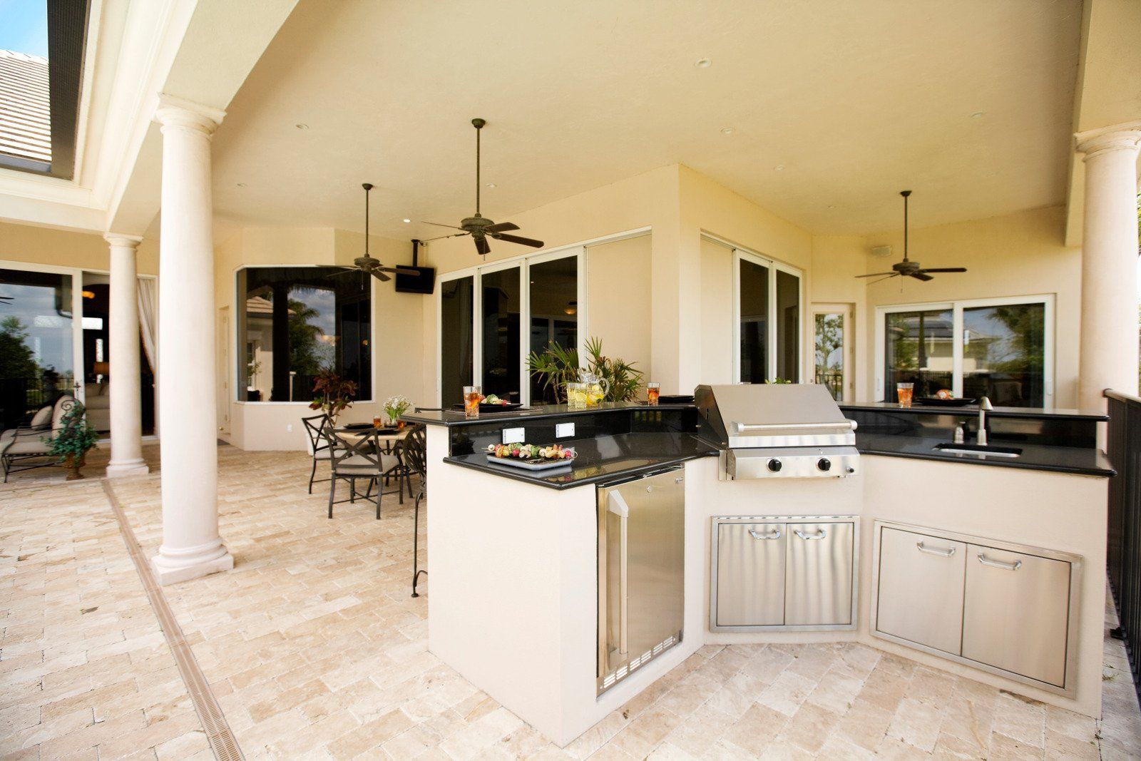 Outdoor kitchen with a grill, sink, and refrigerator; beige tile floor and white columns.