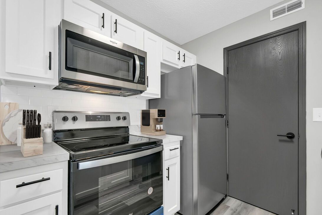 Compact white kitchen with stainless steel stove, microwave, and fridge, next to a gray door
