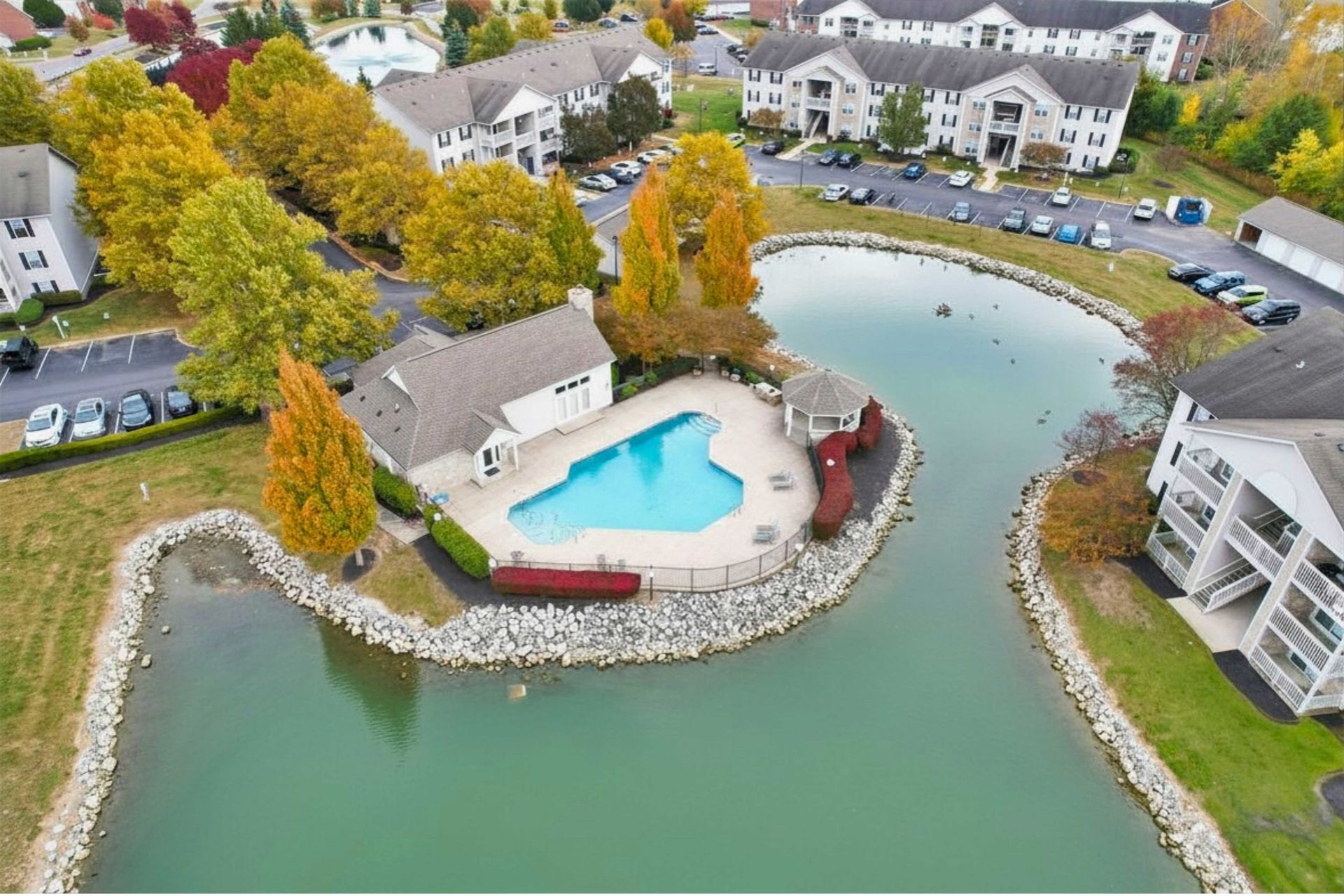 Aerial view of a lakeside apartment complex with a pool, parking lot, and autumn trees