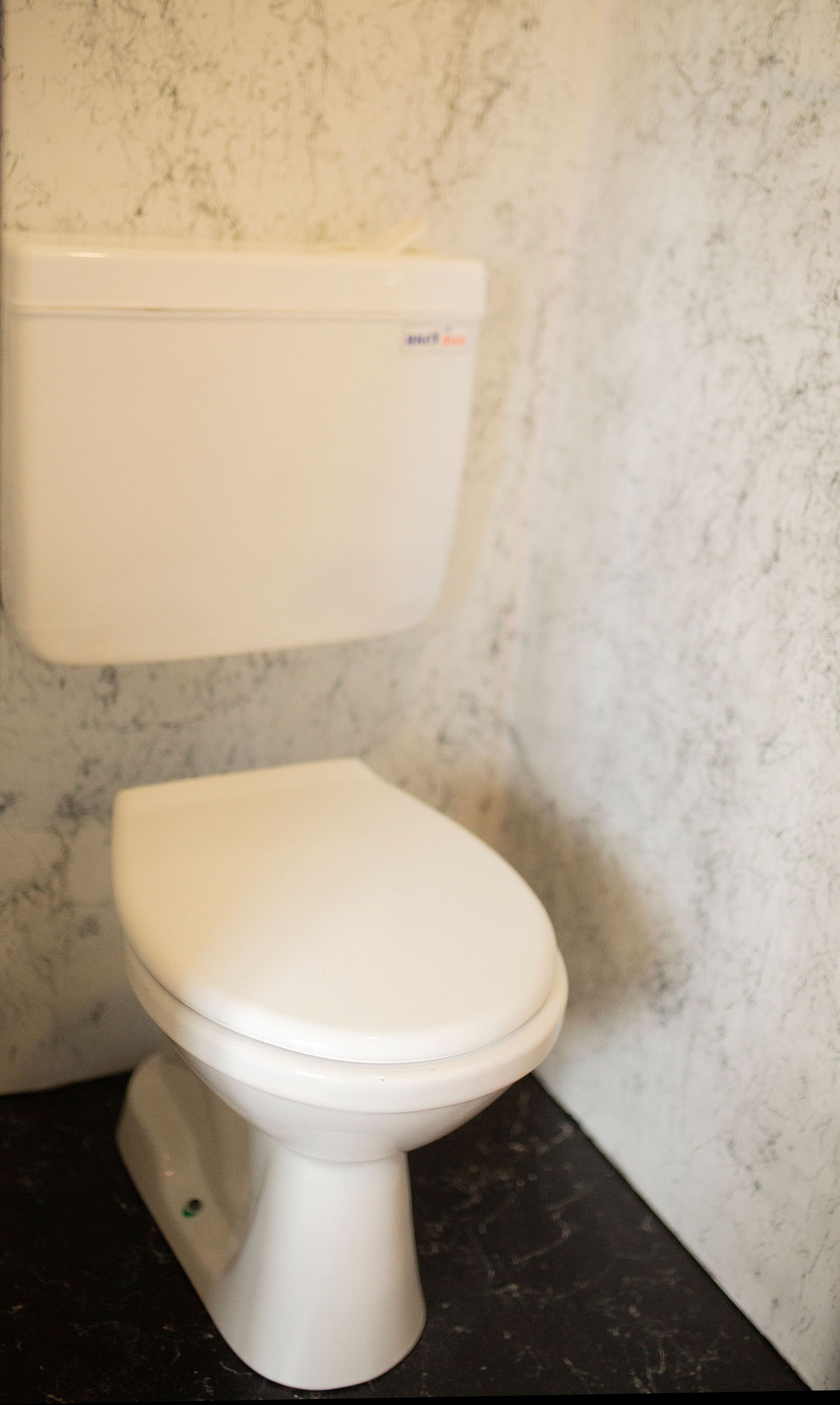 White toilet with tank in a small bathroom with marbled wallpaper and dark floor.