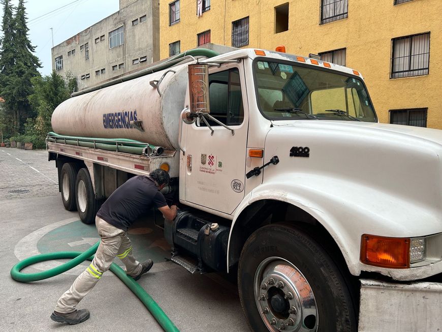 Worker washing a white tanker truck with a green hose on a city street