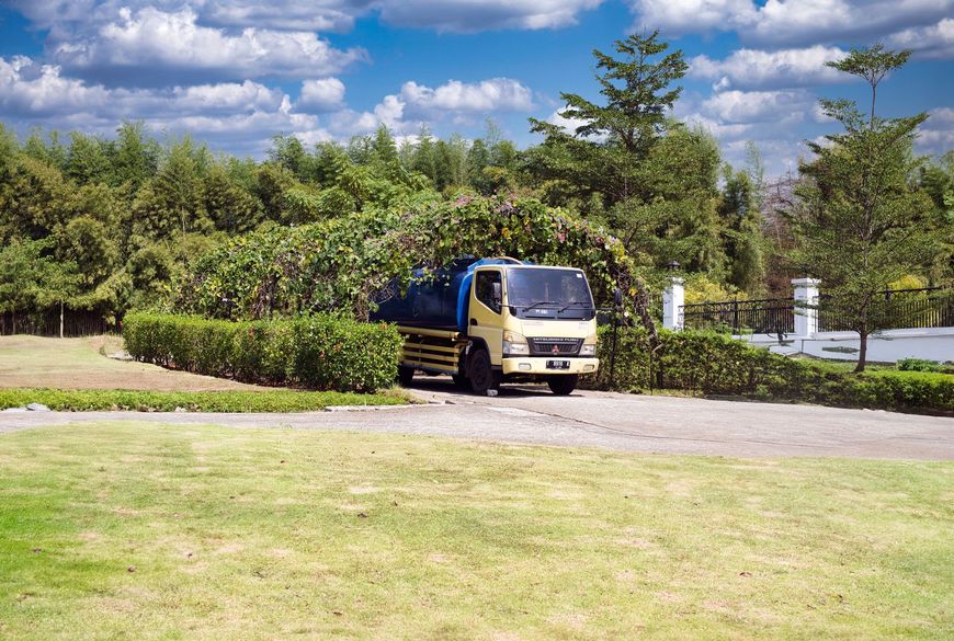 Yellow delivery van driving on a paved road beside green lawns and trees under a blue sky