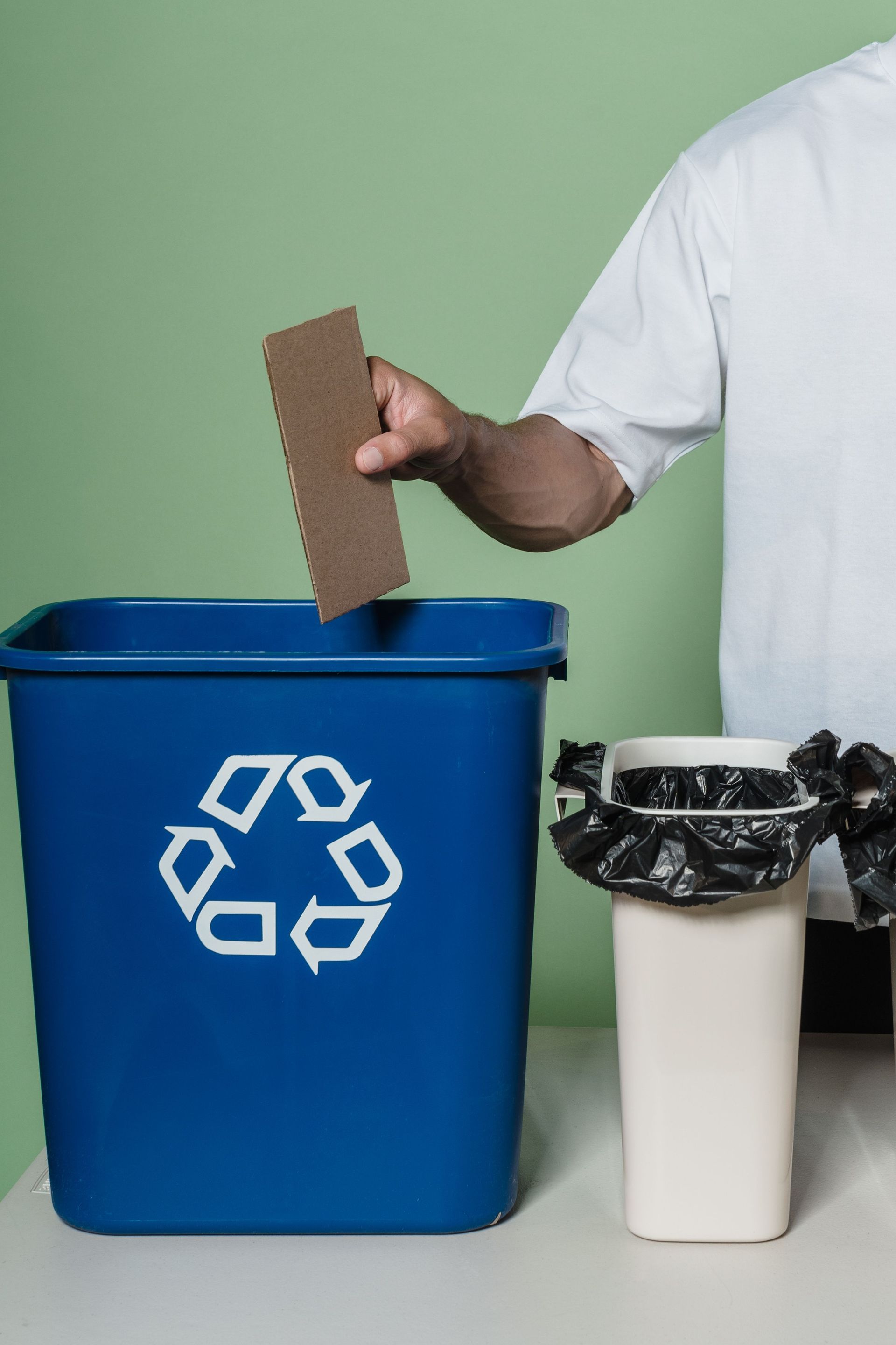 Hand placing cardboard into blue recycling bin beside white trash can