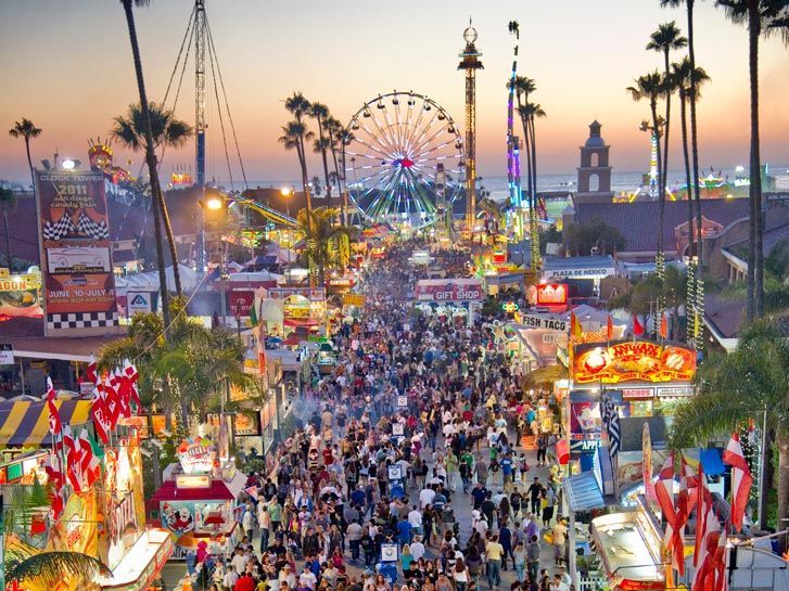A large crowd of people are gathered at a carnival with a ferris wheel in the background.