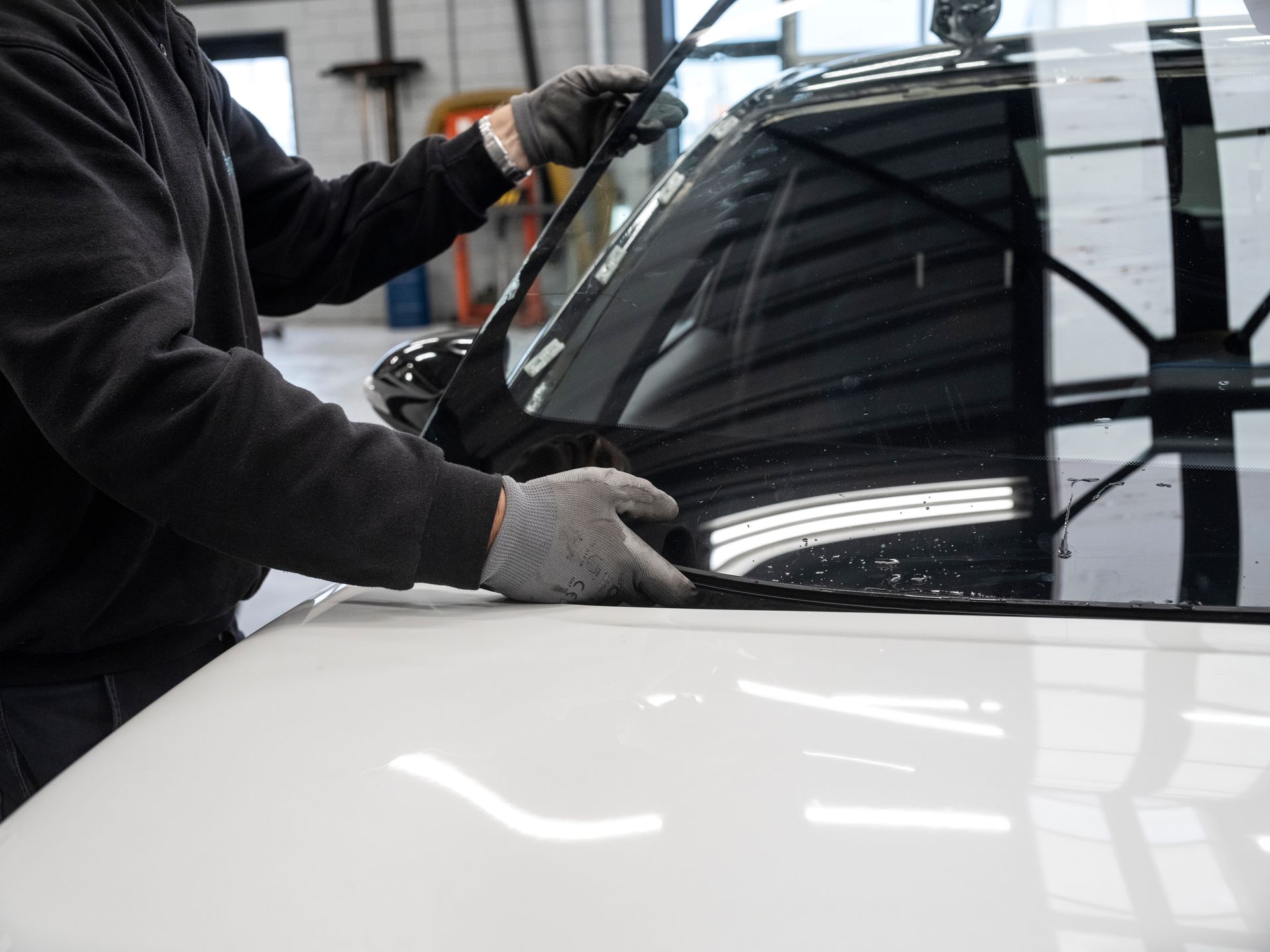 Technician hands performing an auto windshield installation on a white vehicle in a shop.