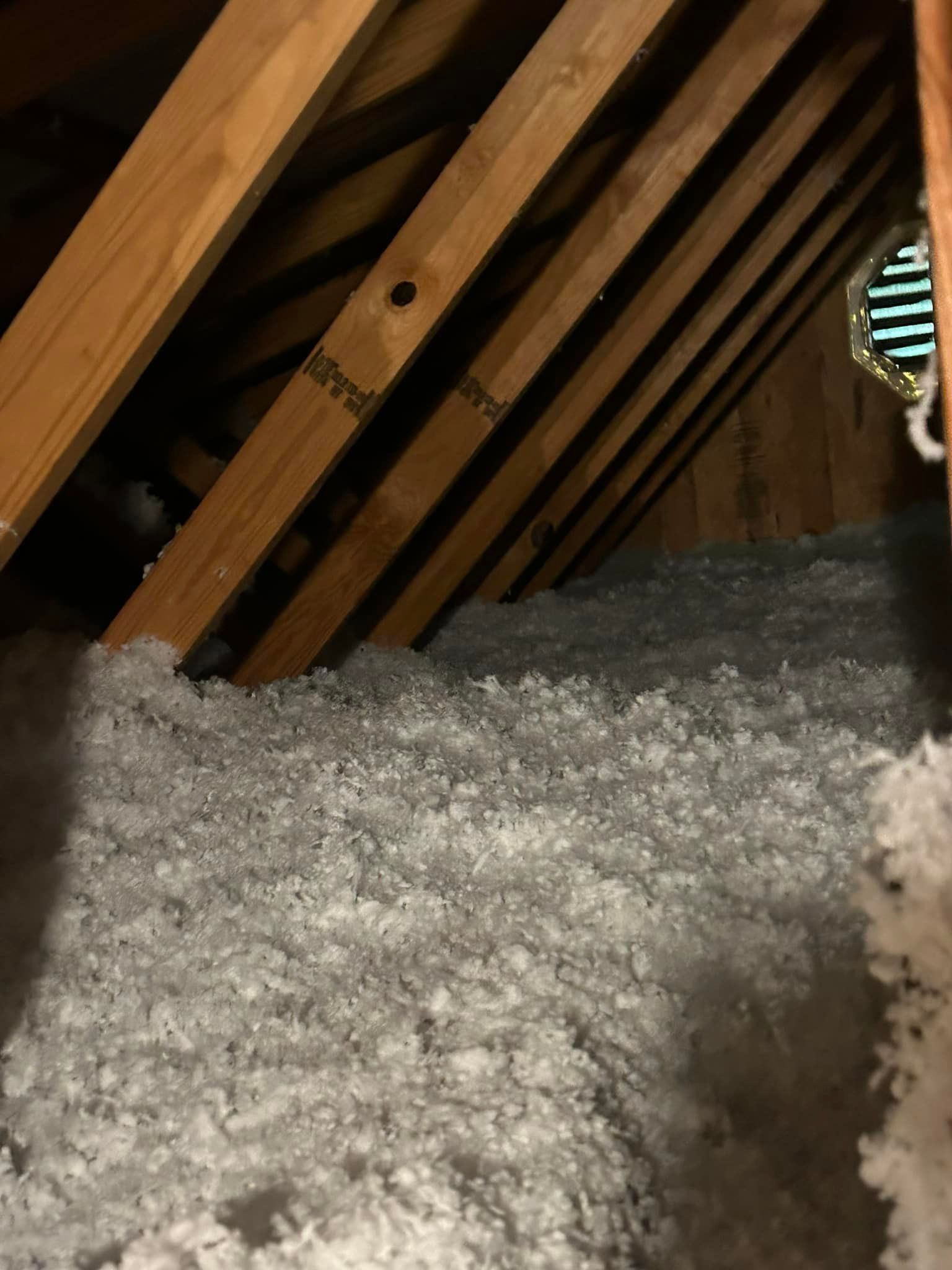Attic interior with angled wooden beams and thick, light gray insulation. Dark shadows and a hint of light from a small opening on the right.