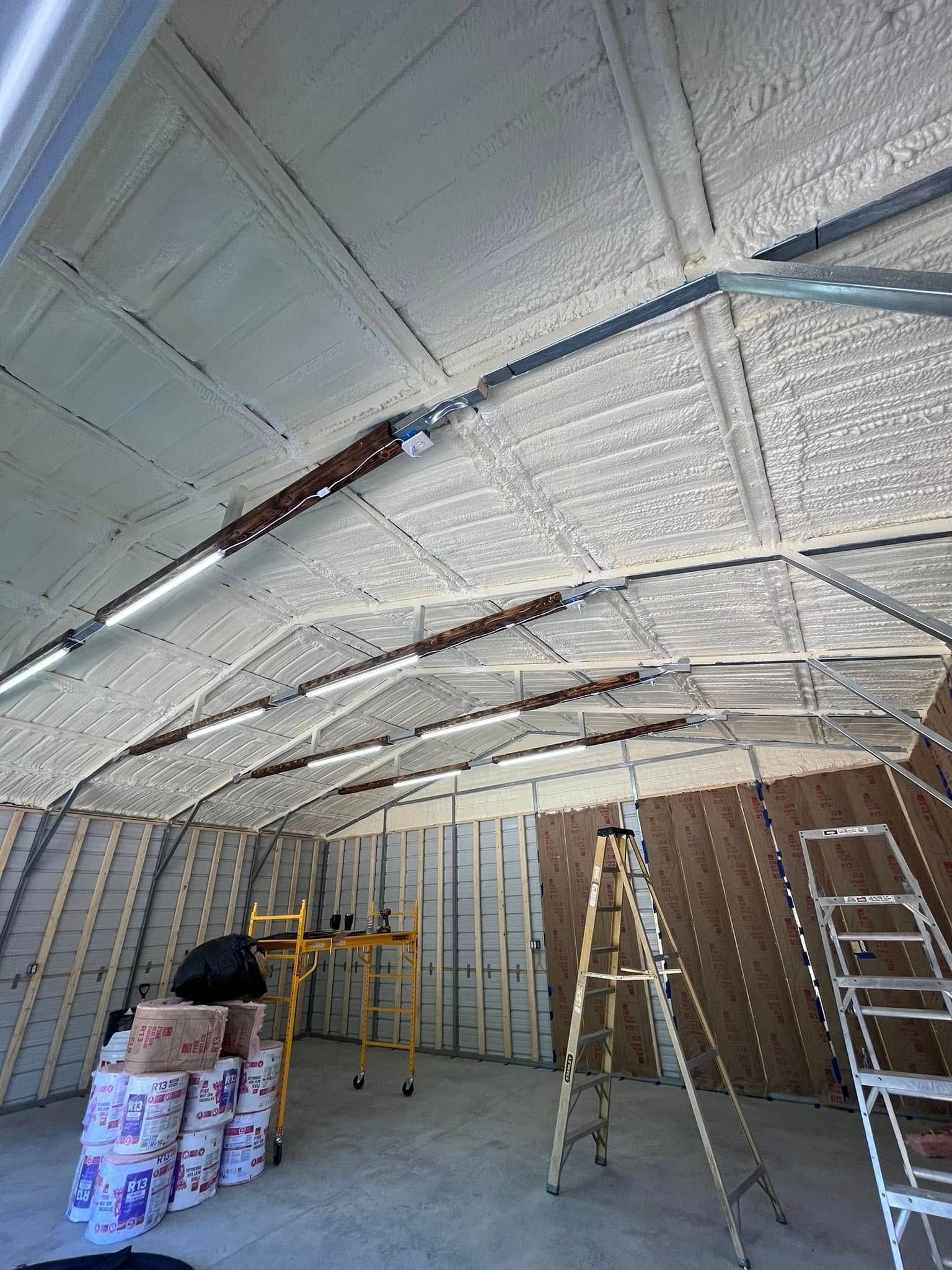 Interior of a metal building with spray foam insulation on the ceiling and walls. Ladders and painting supplies are visible.