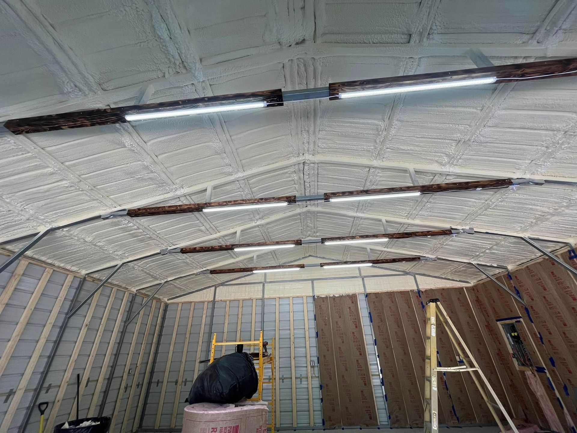 Interior of a metal-framed building with spray foam insulation and LED lights mounted on the arched ceiling. Walls are partially finished with wood panels.