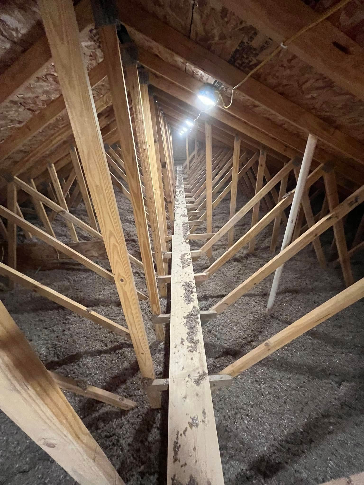 An attic with wooden rafters and insulation. A wooden beam runs down the center, and a light is visible in the distance.