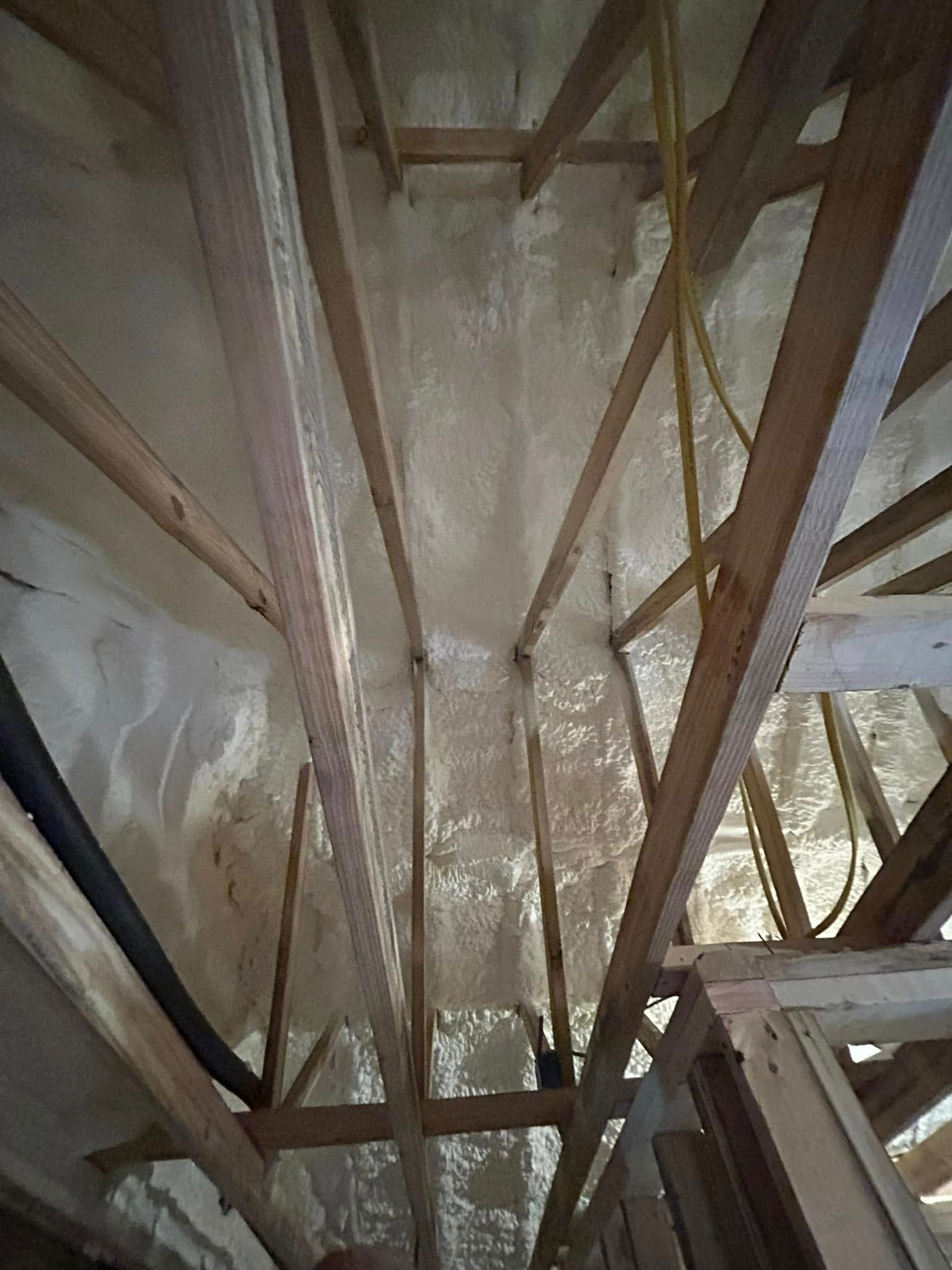 Interior view of an attic space with wooden beams and spray foam insulation. The walls and beams are covered in a white, textured material.