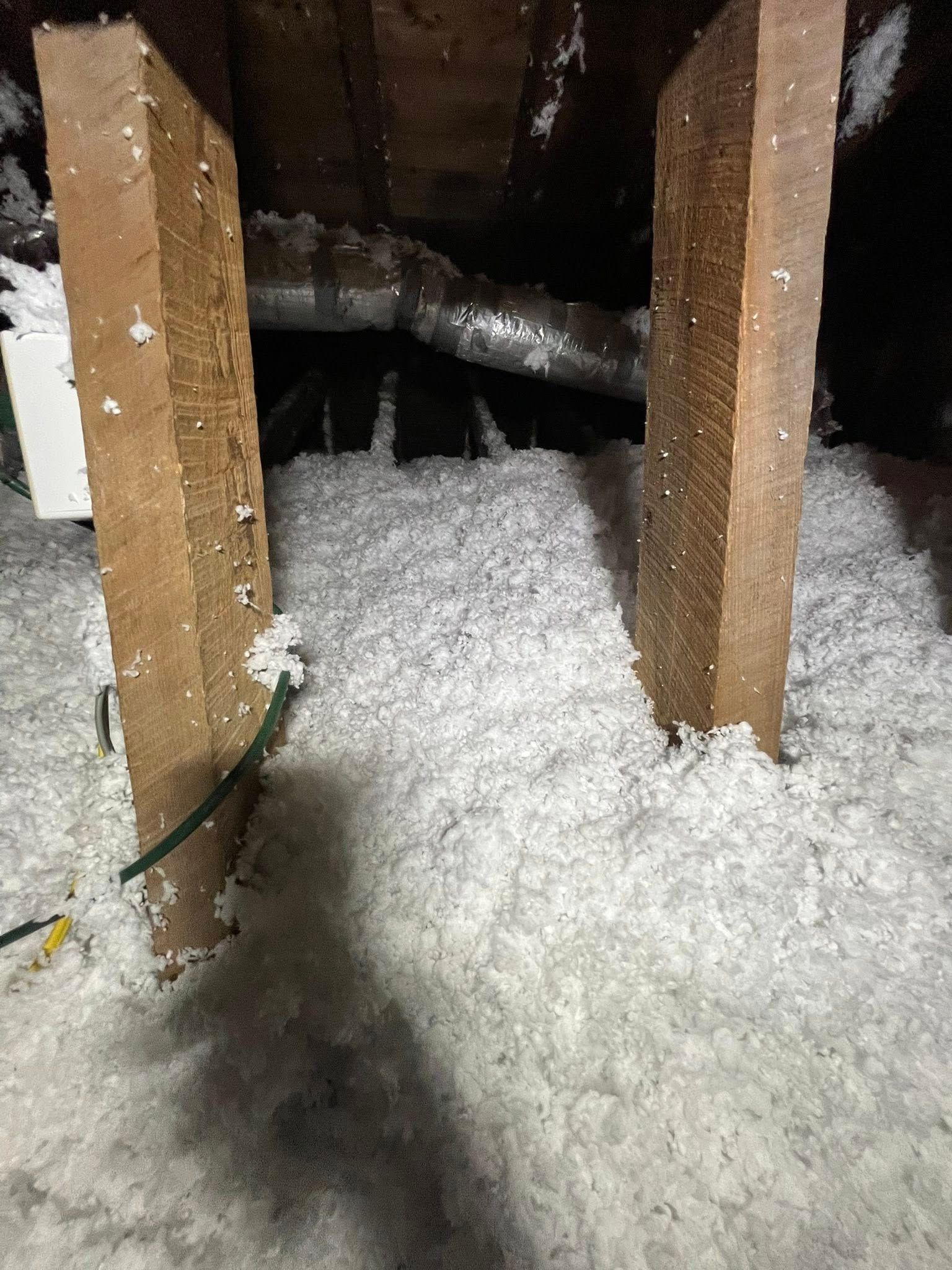 Attic with two vertical wooden beams surrounded by thick, white insulation. A metal duct and wiring are visible in the background.