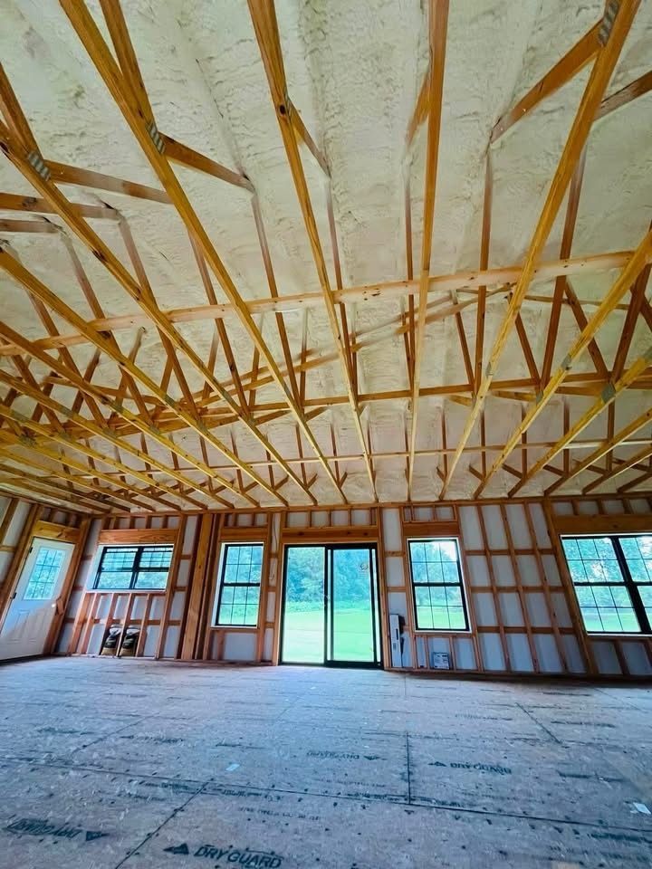 Empty unfinished room with exposed wooden ceiling trusses, framed walls, and large windows letting in daylight