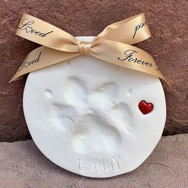 White round memorial plaque with a molded baby handprint, gold ribbon, and a small red heart.
