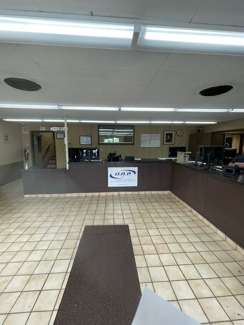 A view of a commercial interior with a service counter, a dark floor mat, and tile flooring under fluorescent lighting.