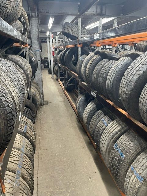 A narrow aisle in a warehouse featuring shelves filled with stacked black rubber tires on both sides.