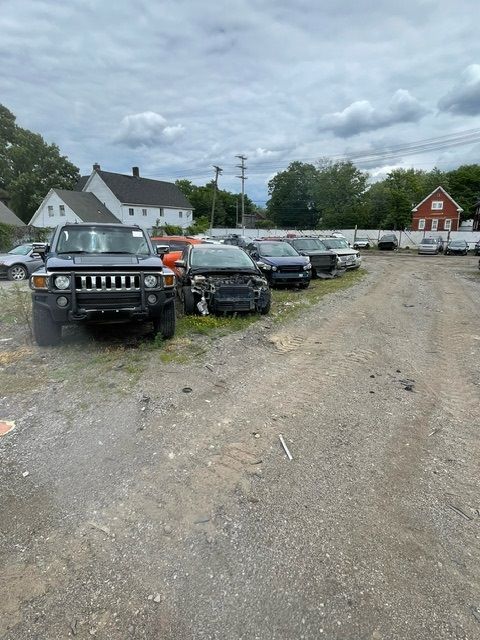 A row of salvaged cars, including a black Hummer, parked on a gravel lot near suburban houses under a cloudy sky.