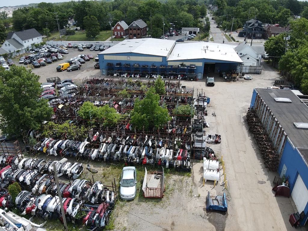 An aerial view of a vehicle salvage yard with rows of wrecked cars, a large blue warehouse, and surrounding greenery.