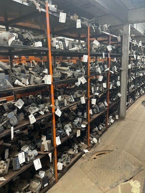 A storage aisle in a warehouse filled with shelves packed with various metal automotive parts, each labeled with white tags.